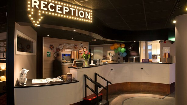 Een receptiegedeelte met een bord waarop 'RECEPTION' staat, een balie met een beeldje van een hond, en een trap naar een hoger niveau.