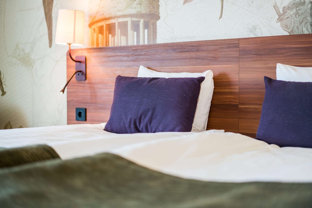 Een netjes opgemaakt bed met een houten hoofdbord, twee kussens en een wandlamp.