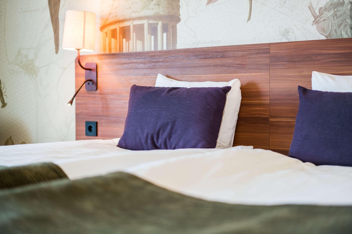 Un lit bien fait, avec une literie blanche et deux oreillers bleu foncé, une tête de lit en bois et une lampe fixée au mur.