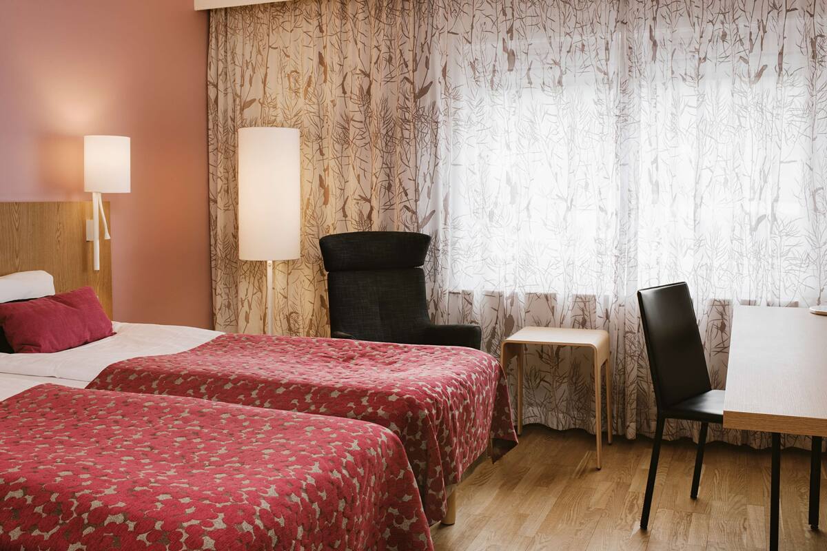 Camera d'albergo con due letti, una scrivania, una sedia e una lampada.