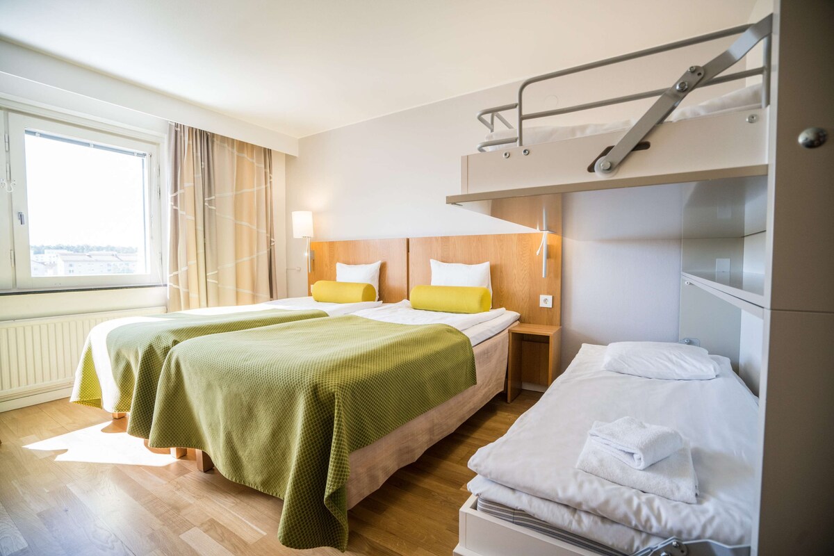 Habitación de hotel con dos camas, una litera, piso de madera, ventana con cortinas y una lámpara de noche.