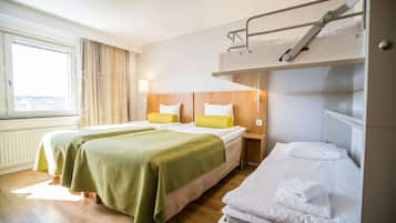 A hotel room with two beds, a bunk bed, wooden flooring, a window with curtains, and a bedside lamp.