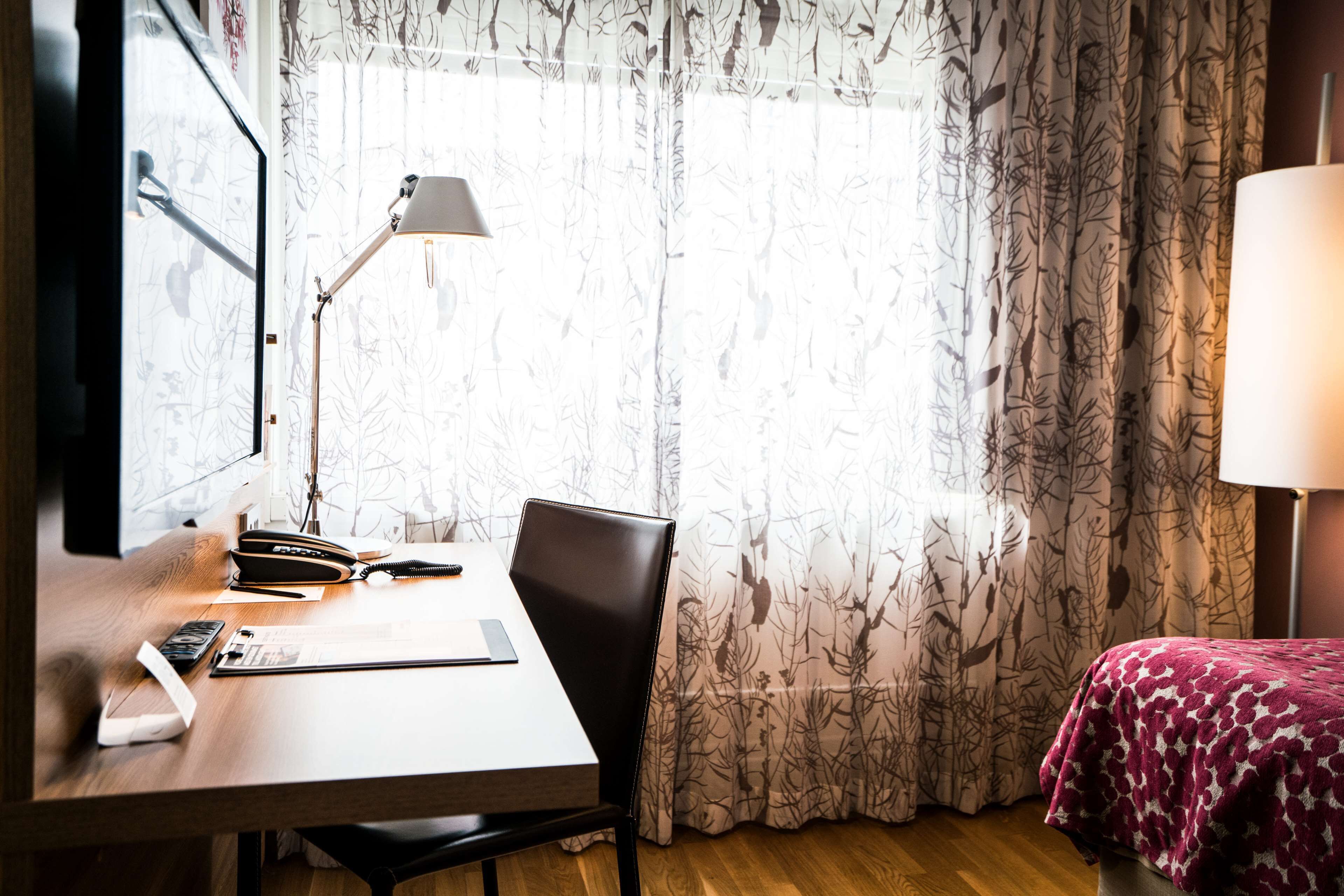 A hotel room with a wooden desk, a chair, a bed with a patterned blanket, a window with sheer curtains, and a standing lamp.