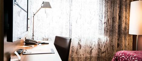 A hotel room with a wooden desk, a chair, a bed with a patterned blanket, a window with sheer curtains, and a standing lamp.