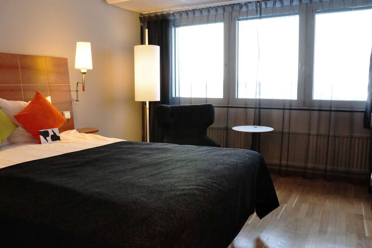 Habitación de hotel con una cama grande, una mesita de noche, una silla, una pequeña mesa redonda y una ventana con cortinas.