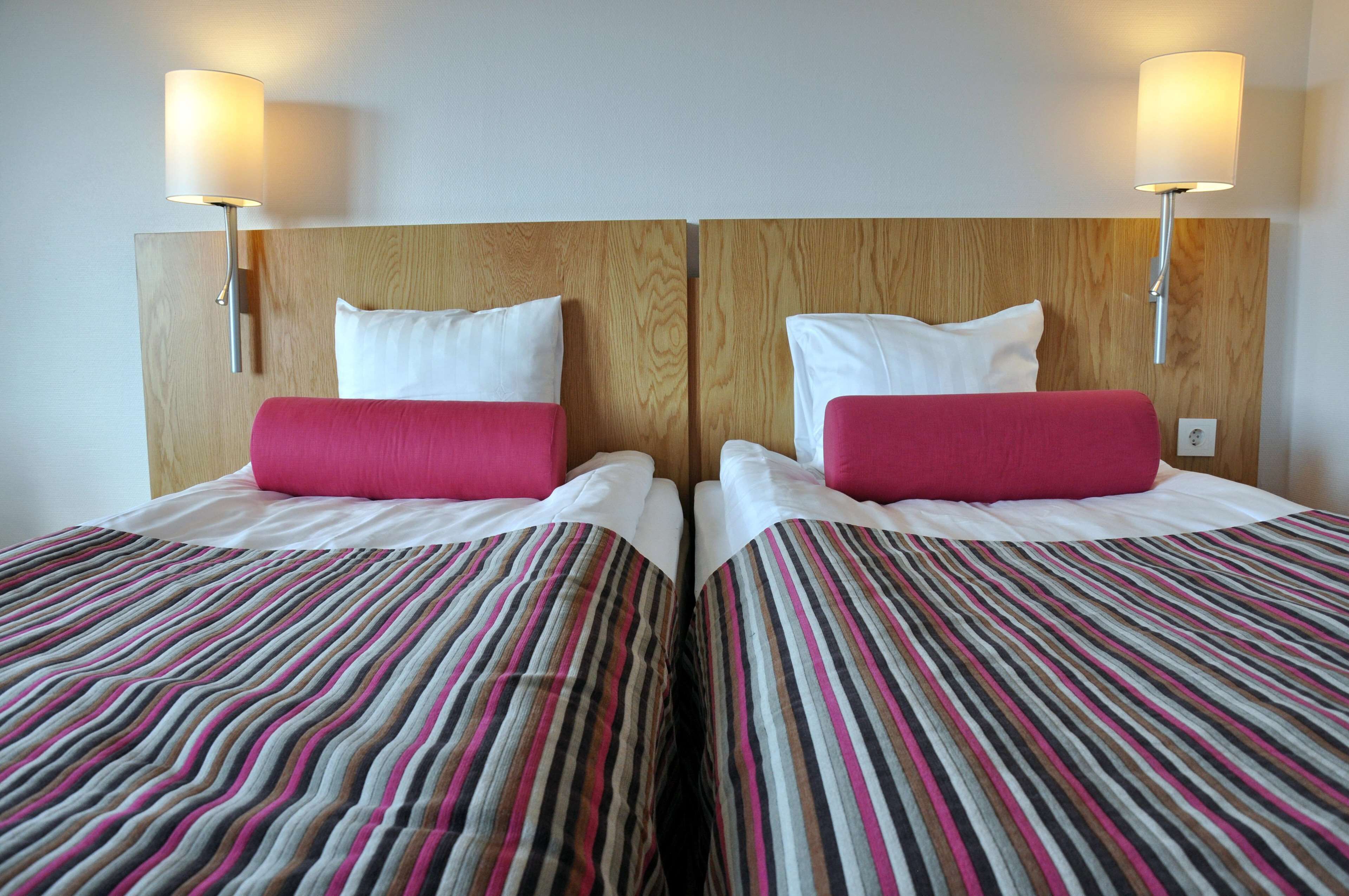 A hotel room with two beds, each with a wooden headboard, white bedding, and a striped blanket. There are wall-mounted lamps on either side of the beds.