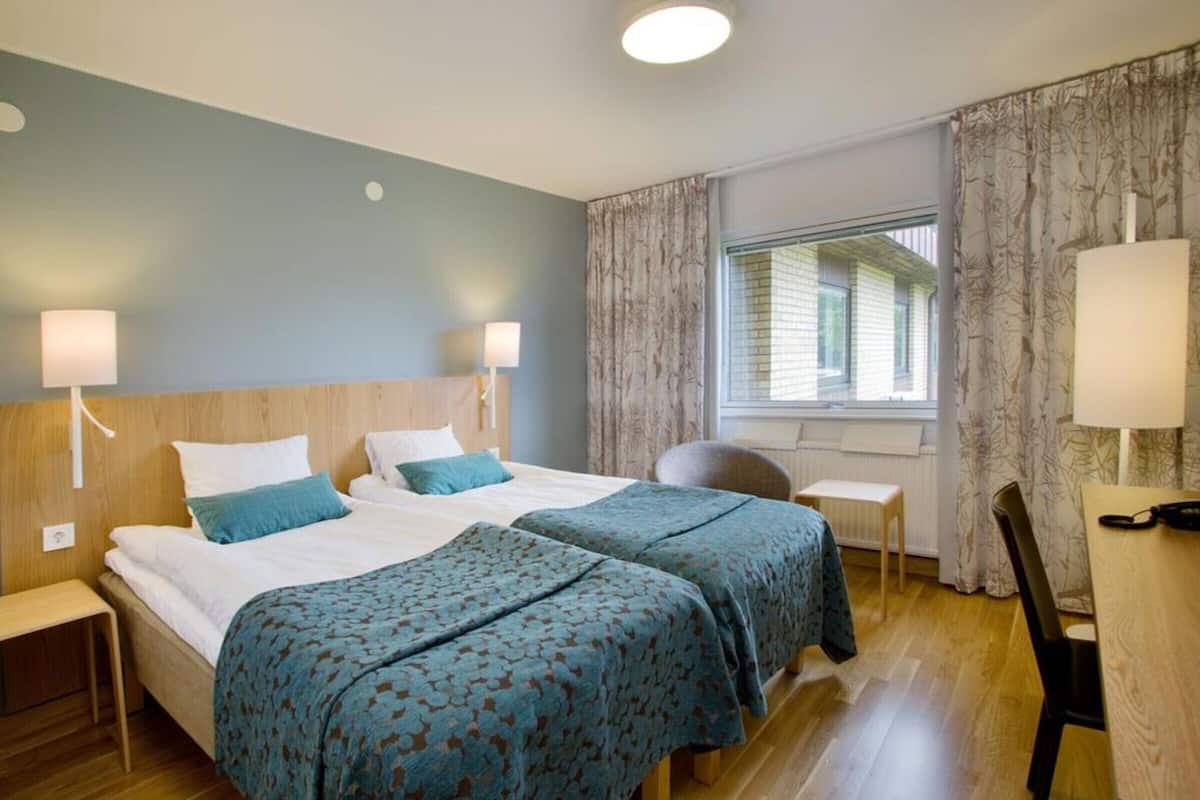 A hotel room with two beds, a desk, a chair, and a window with curtains.