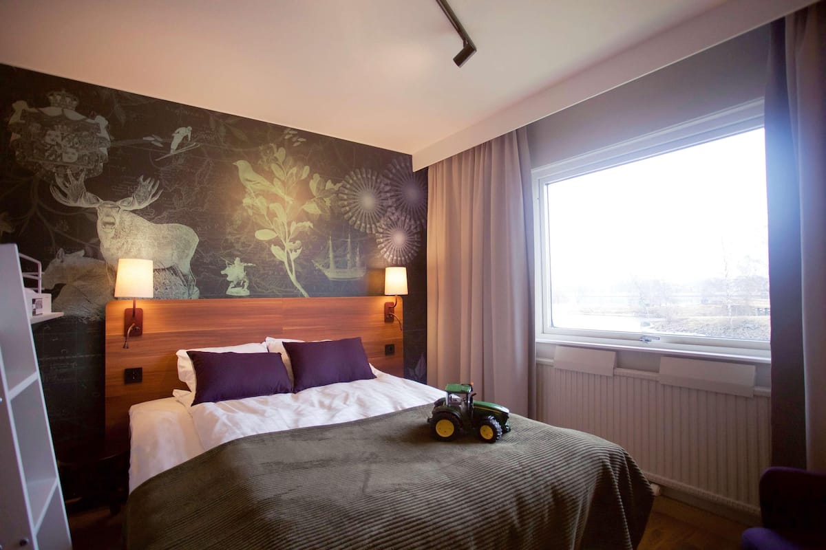 A hotel room with a large bed, two bedside lamps, a mural on the wall, and a window with a view.