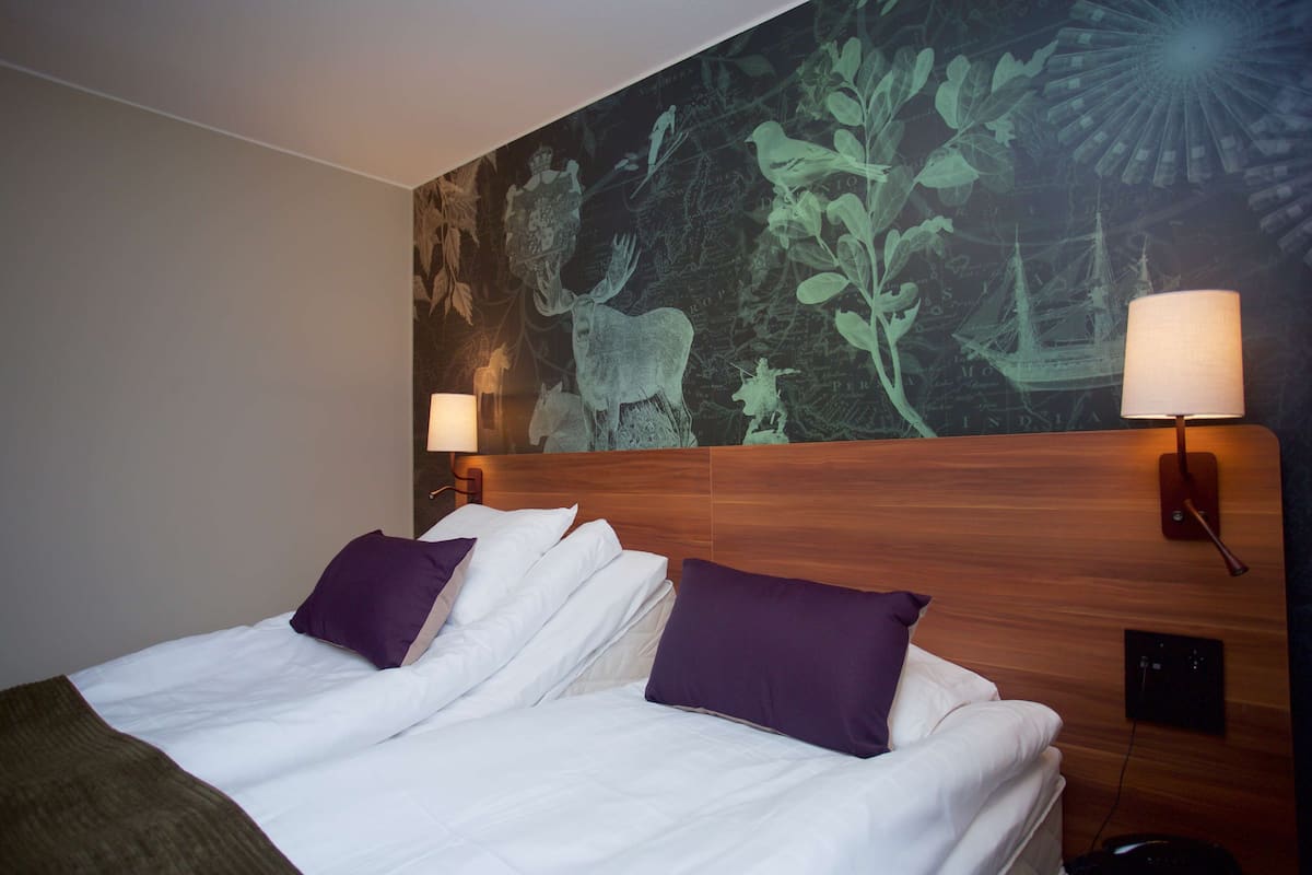 A hotel room with a large bed, two purple pillows, a wooden headboard, and a wall mural.