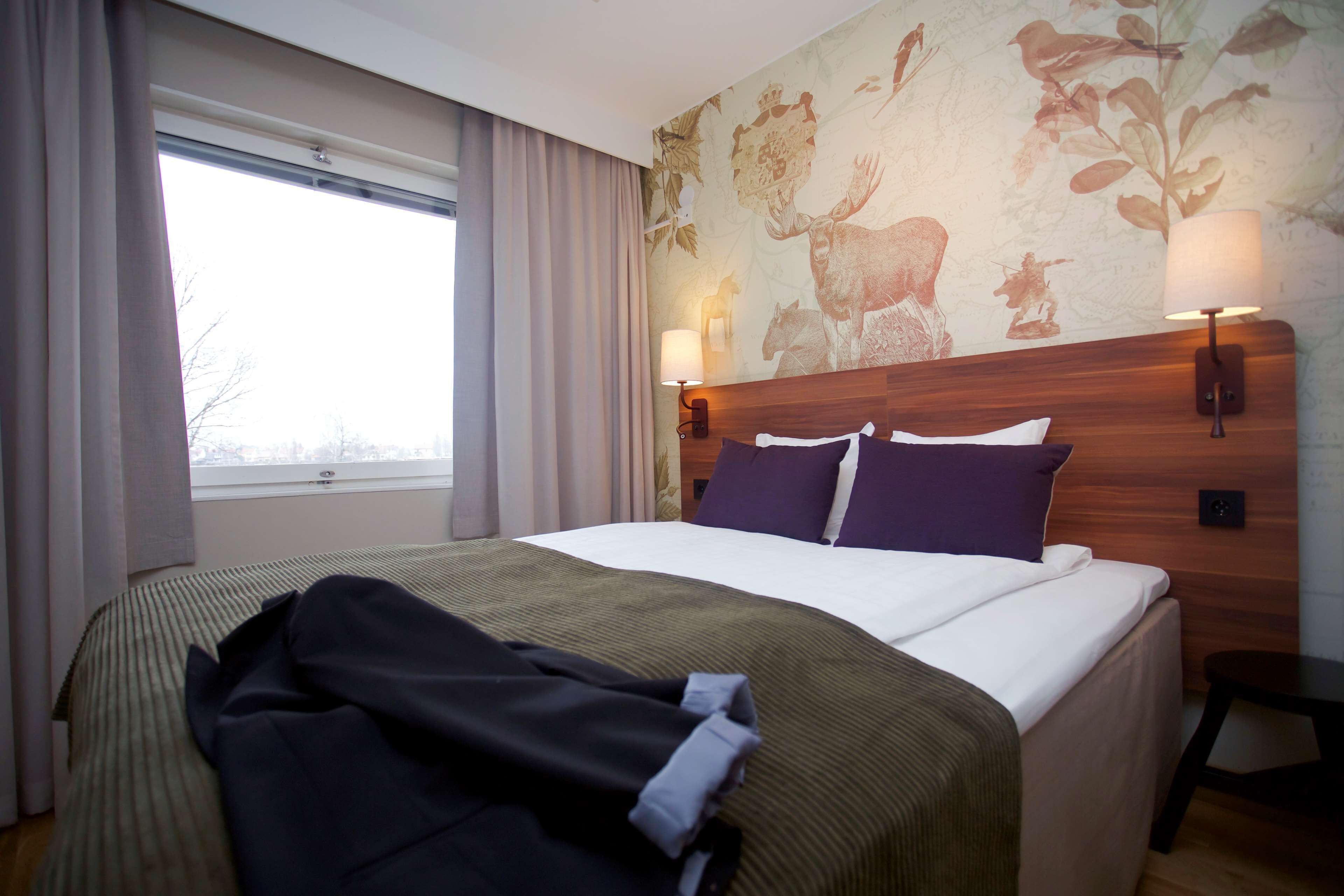 A hotel room with a large bed, a window with curtains, a wall with a floral mural, and two wall-mounted lamps.