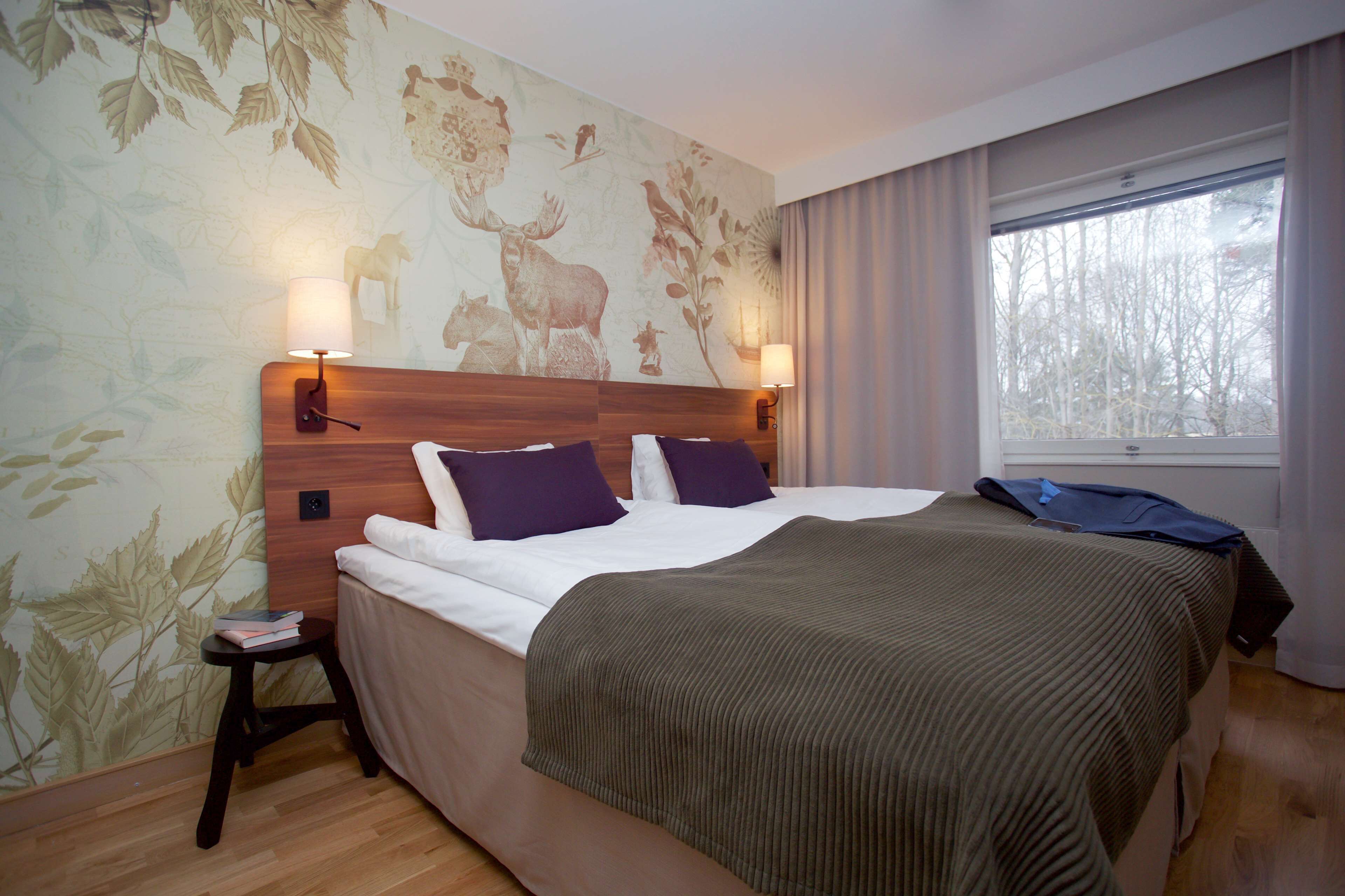 Habitación de hotel con una cama grande, dos lámparas de pared, una mesita de noche pequeña y una ventana con cortinas.