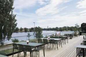 An outdoor dining area with tables and chairs overlooking a lake, with a view of a distant building. - Scandic Klarälven (Karlstad)