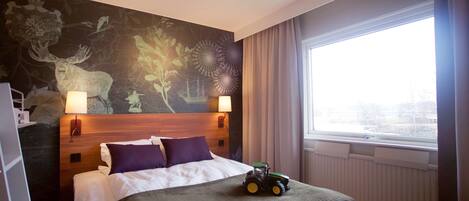 A hotel room with a large bed, a mural on the wall, a window with curtains, and a toy tractor on the bed.