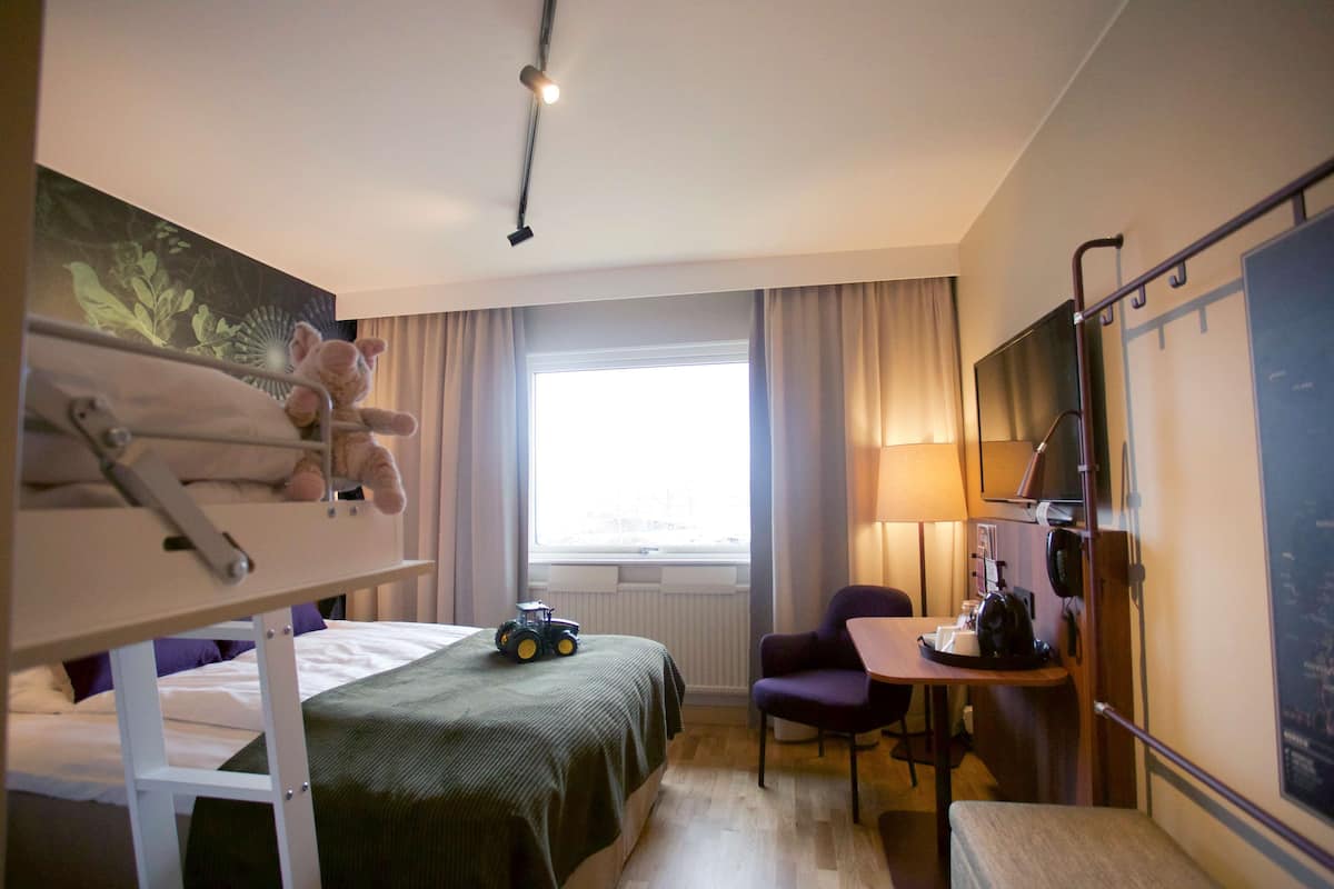 A hotel room with a bunk bed, a desk with a chair, a lamp, and a window with curtains.