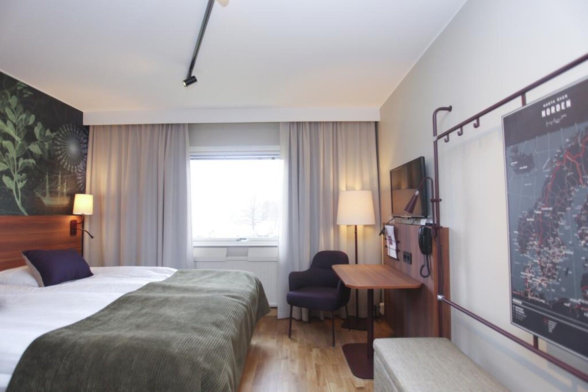 A hotel room with two beds, a desk, a chair, a map on the wall, and a window with curtains.