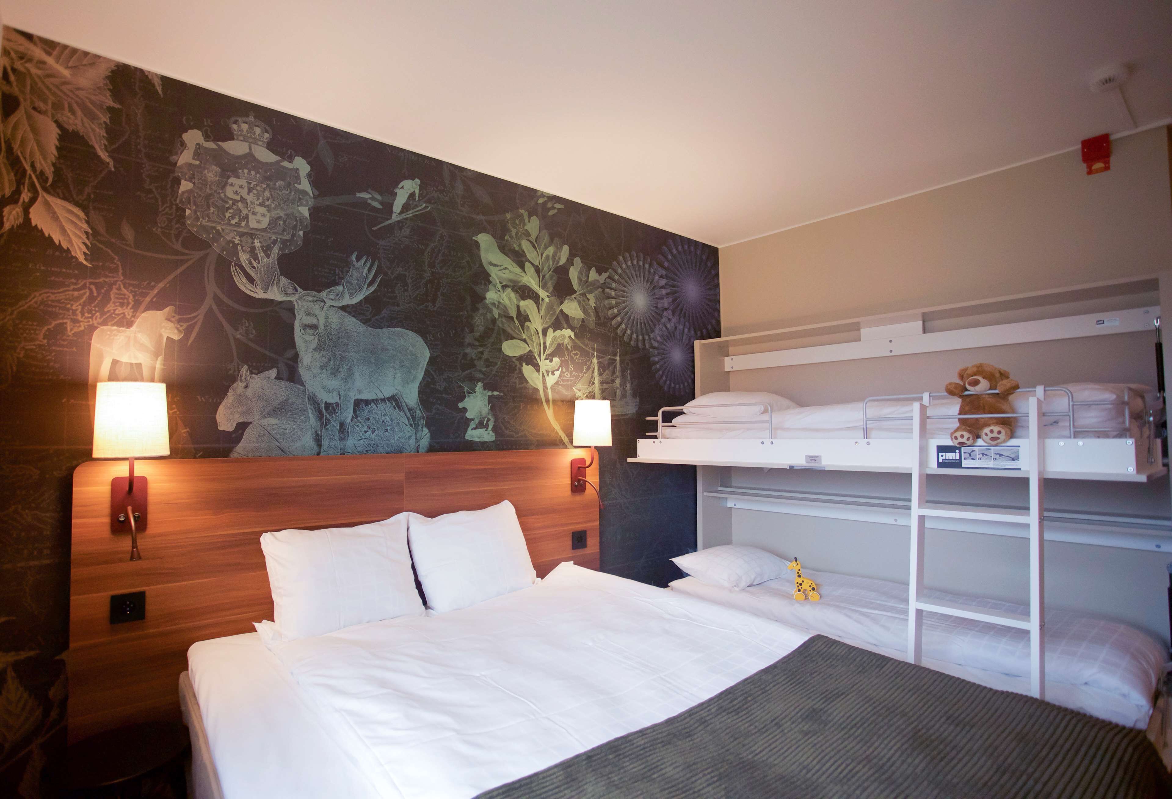 Een hotelkamer met een groot bed, een stapelbed, een muurschildering met bos- en dierdesigns, en twee wandlampen.