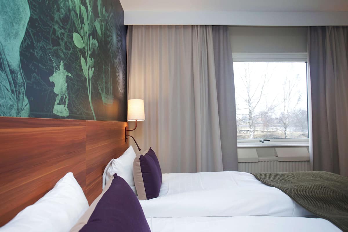 A hotel room with a large bed, a headboard with a mural, a window with curtains, and a wall-mounted lamp.