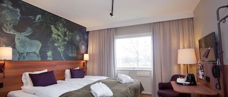 Habitación de hotel con una cama grande, dos mesitas de noche, un escritorio y una silla. Se aprecia una ventana con cortinas, un televisor montado en la pared y un teléfono.