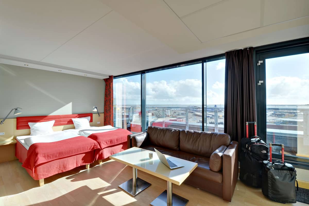A hotel room with two beds, a sofa, a table with a laptop, and large windows with a city view.