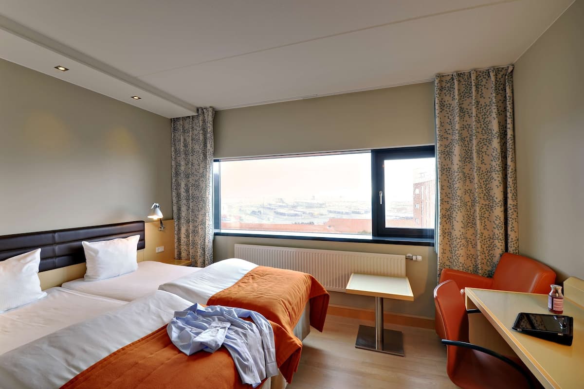 A hotel room with two beds, a small table, a chair, a window with a view, and a radiator.