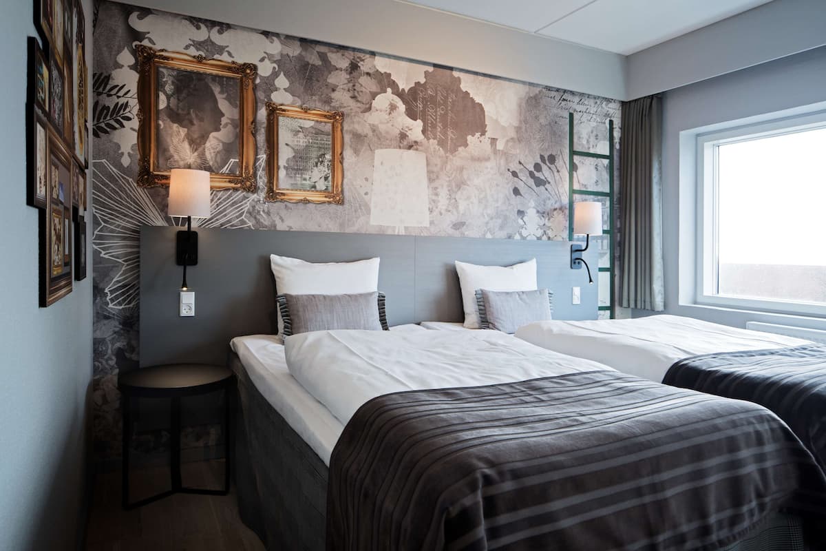 A hotel room with two beds, a small stool, and a large window.