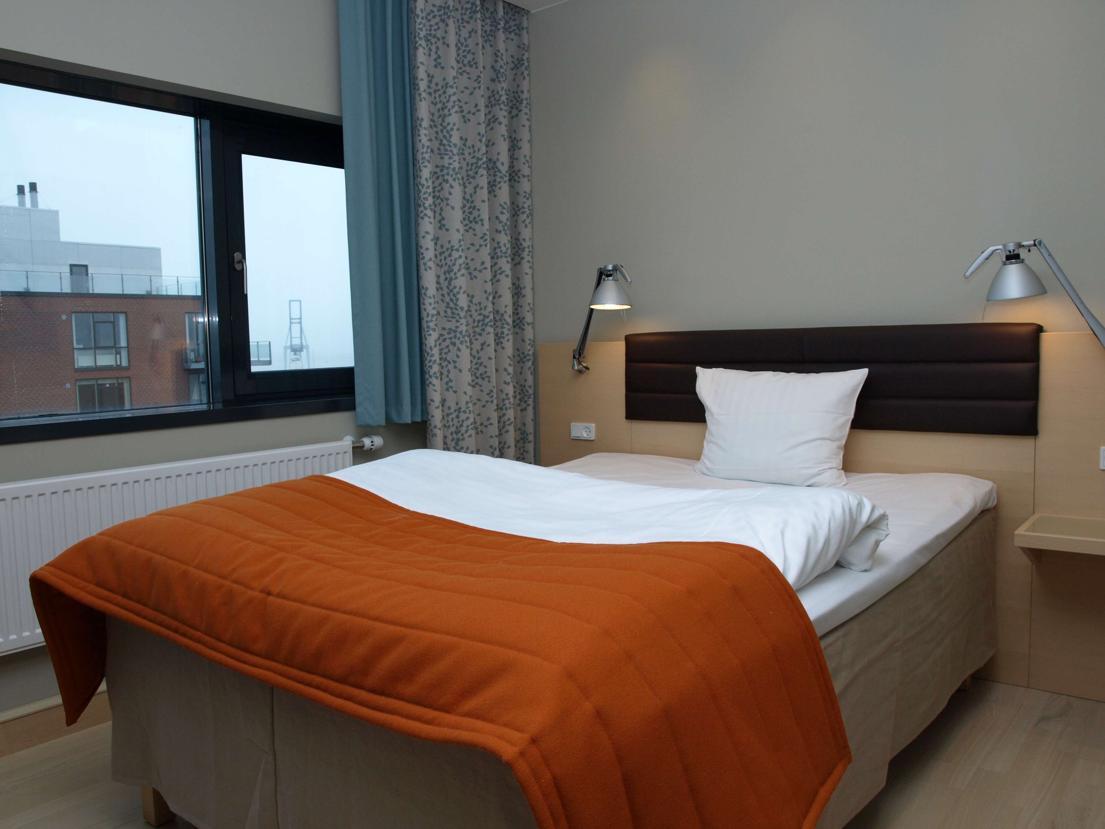 A hotel room with a bed, two bedside lamps, a window with curtains, and a view of buildings outside.