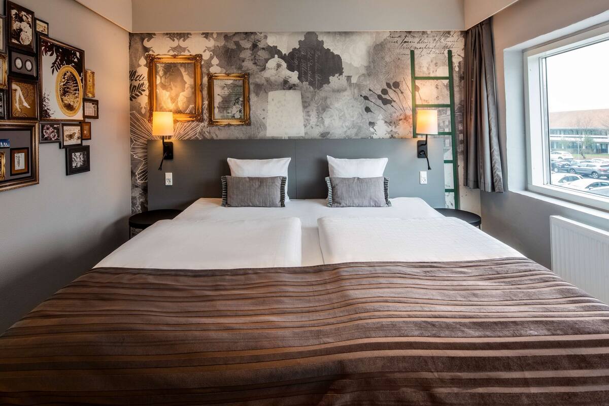 A hotel room with a large bed, two bedside tables, a wall-mounted lamp, and a view of parked cars through the window.