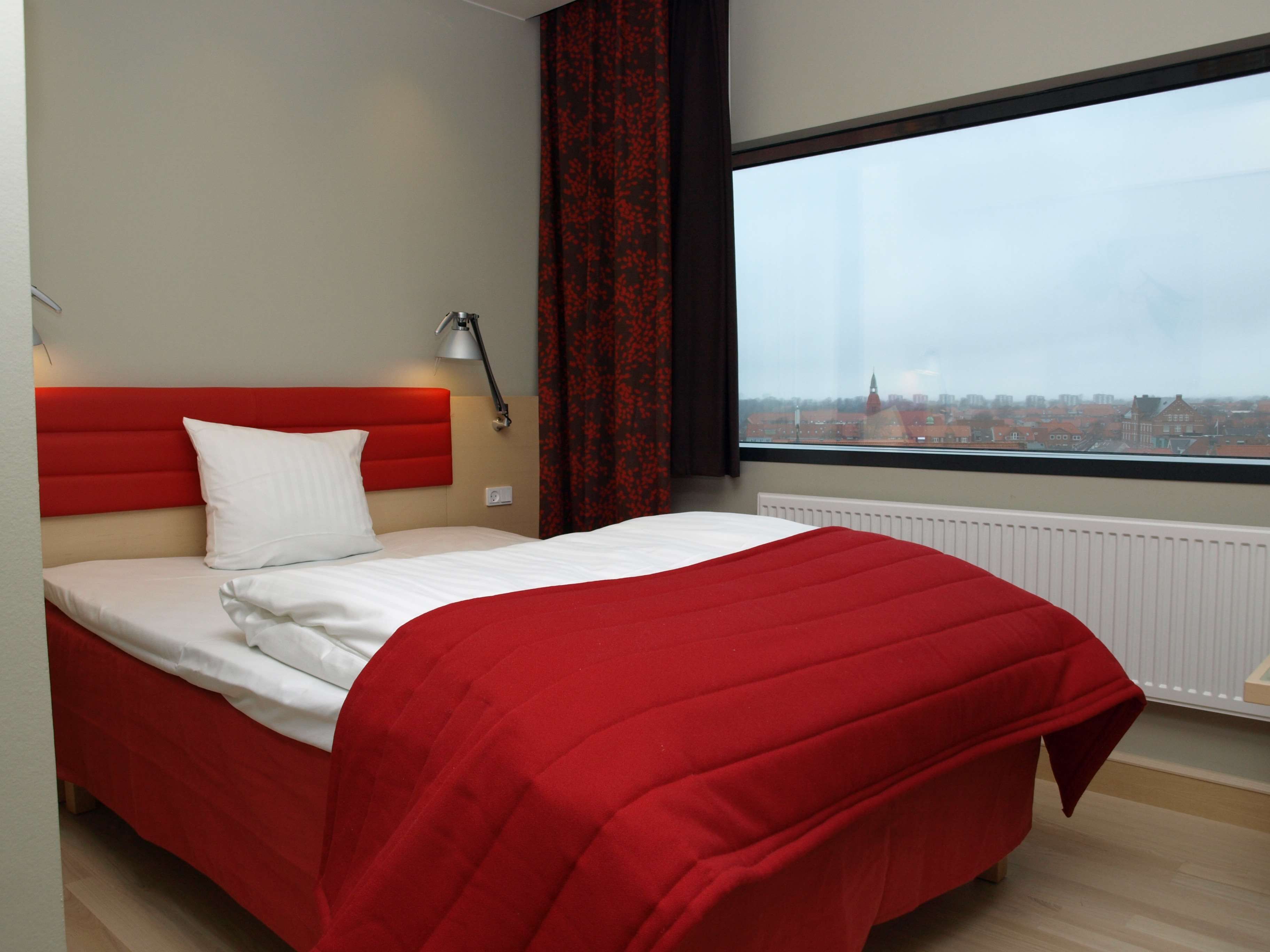 Une chambre d’hôtel moderne avec un grand lit, une tête de lit rouge, une fenêtre donnant sur la ville et un rideau rouge.