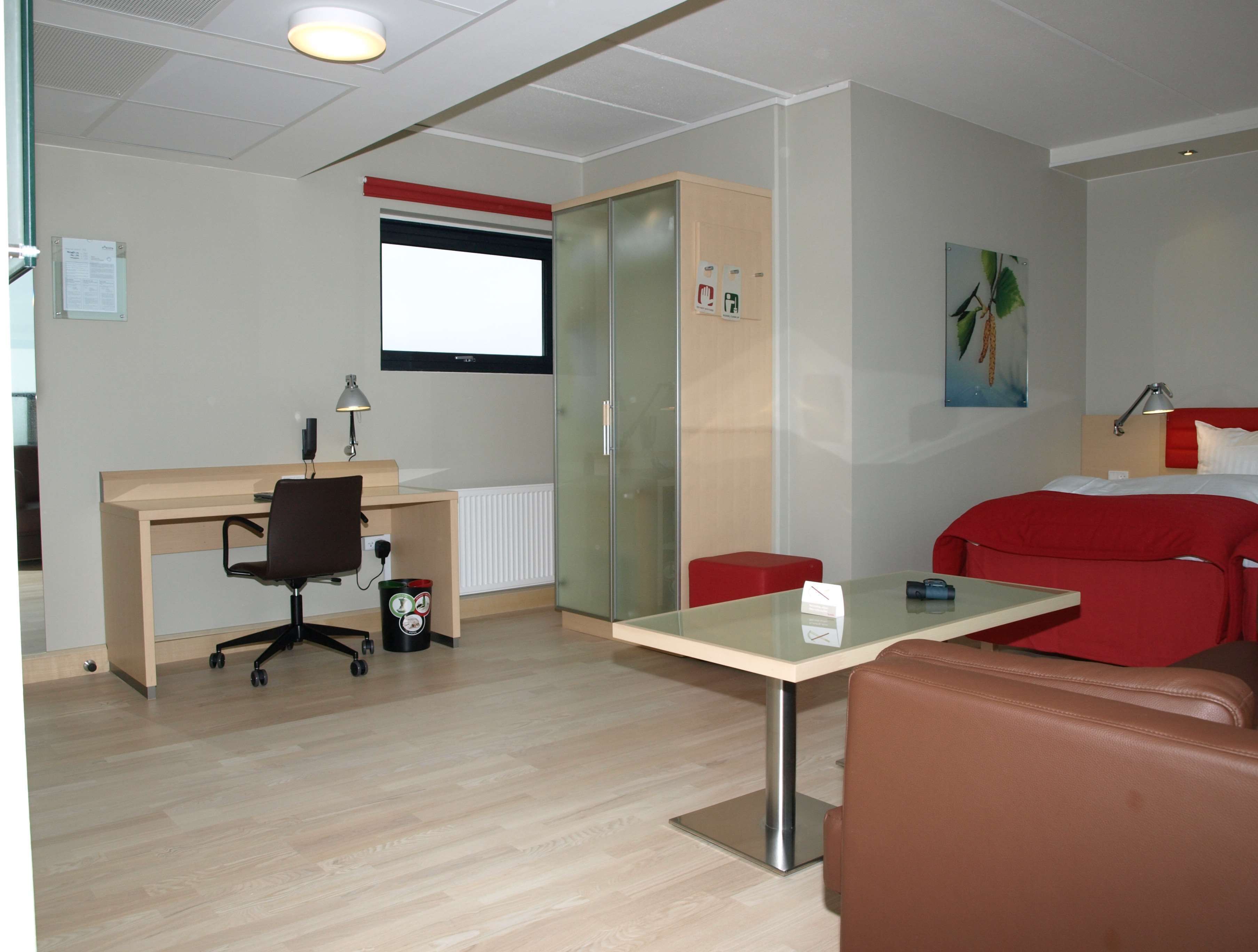 Una habitación de hotel moderna con un escritorio de madera, un sofá rojo, una mesa de vidrio y una silla roja.