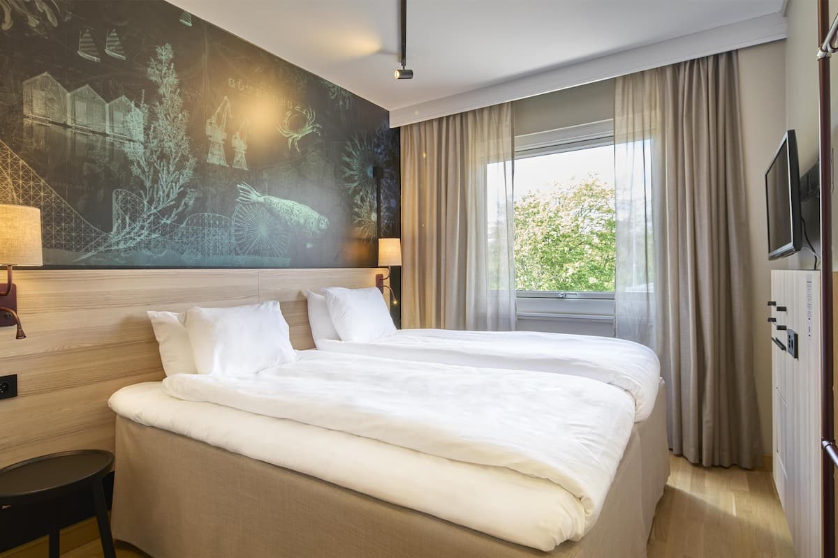 Ett hotellrum med en stor säng, två sängbord, en TV som är monterad på väggen och ett fönster med gardiner.