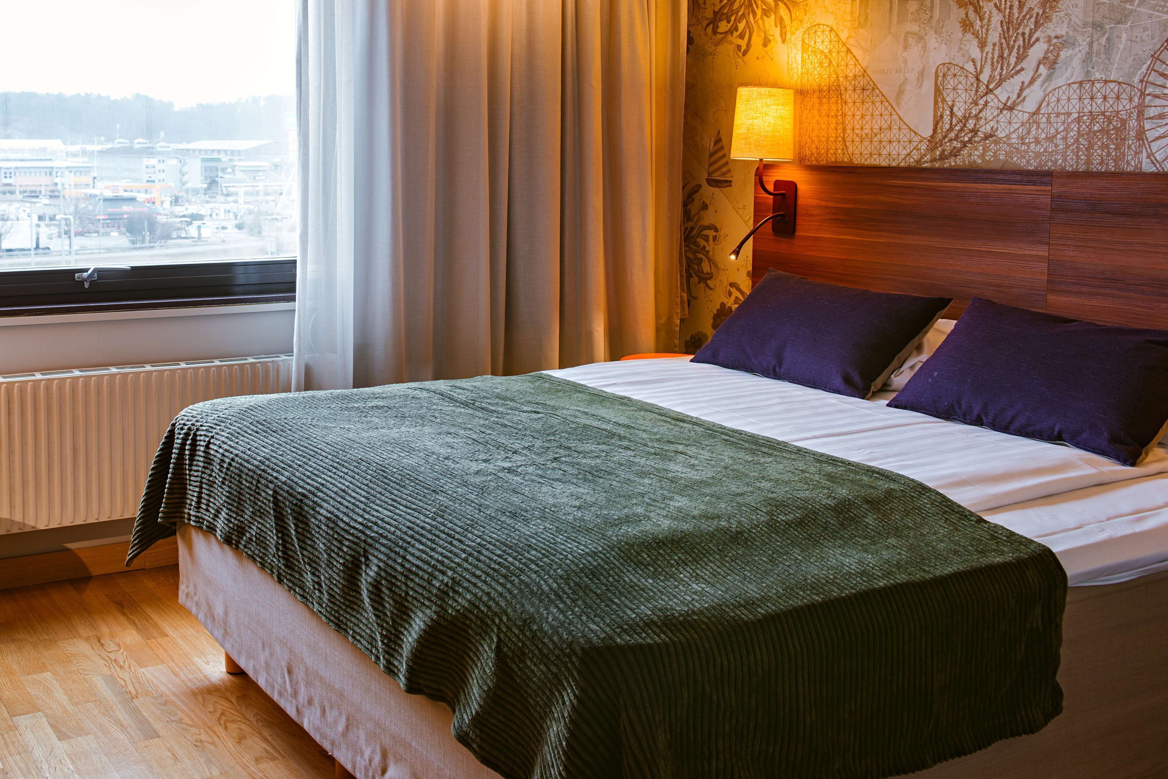 A hotel room with a bed, a window with curtains, a wall-mounted lamp, and a view of the city.