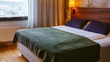 A hotel room with a bed, a window with curtains, a wall-mounted lamp, and a view of the city.
