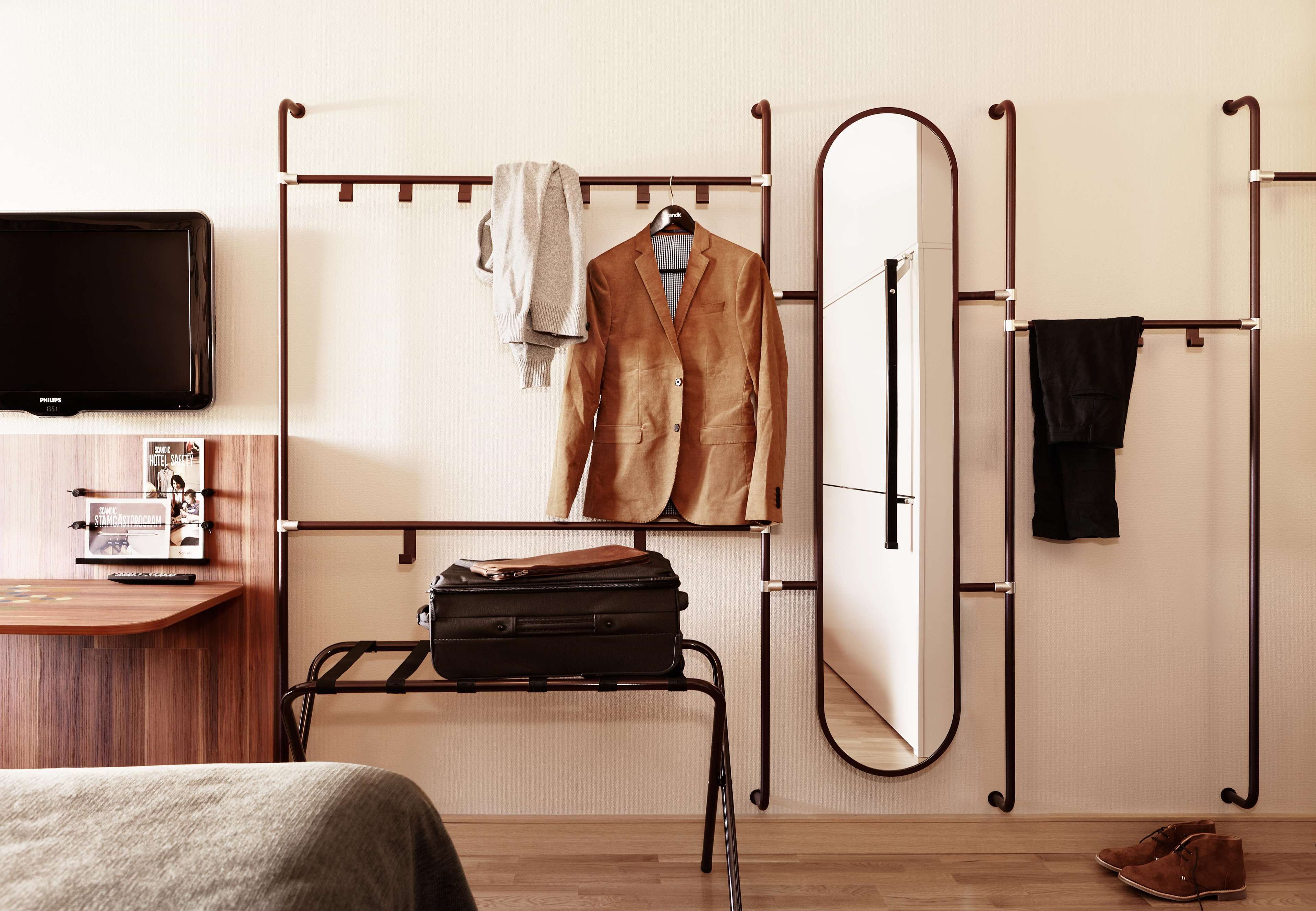 A bedroom with a bed, a bedside table with a remote control, a mirror, a coat rack, and a suitcase.