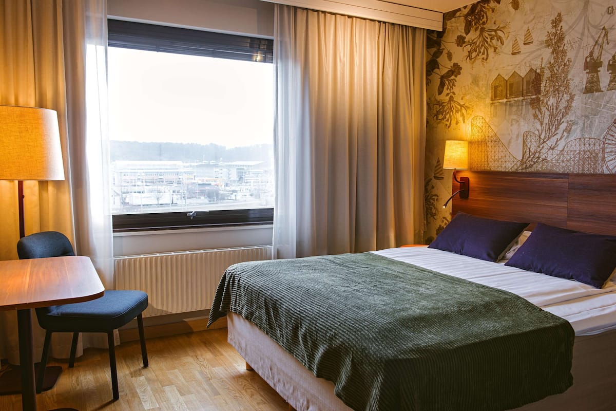 Ett hotellrum med en säng, ett bord, en stol, ett fönster med gardiner och en vägg med en väggmålning.