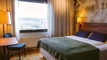 Habitación de hotel con cama, una mesa, una silla, ventana con cortinas y una pared con un mural.