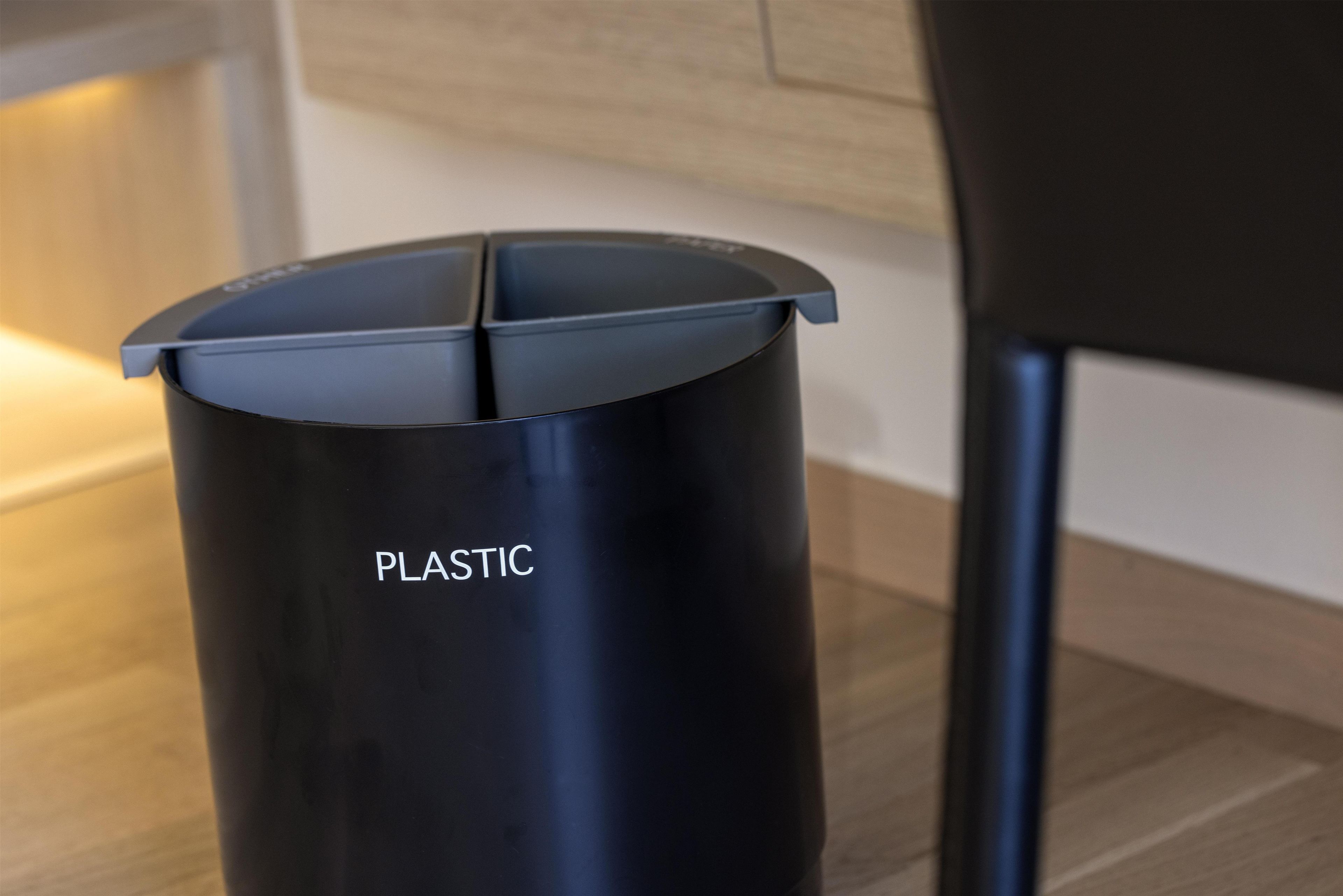 Une poubelle noire en plastique, sur laquelle est inscrit le mot « PLASTIQUE », est posée sur un plancher en bois.