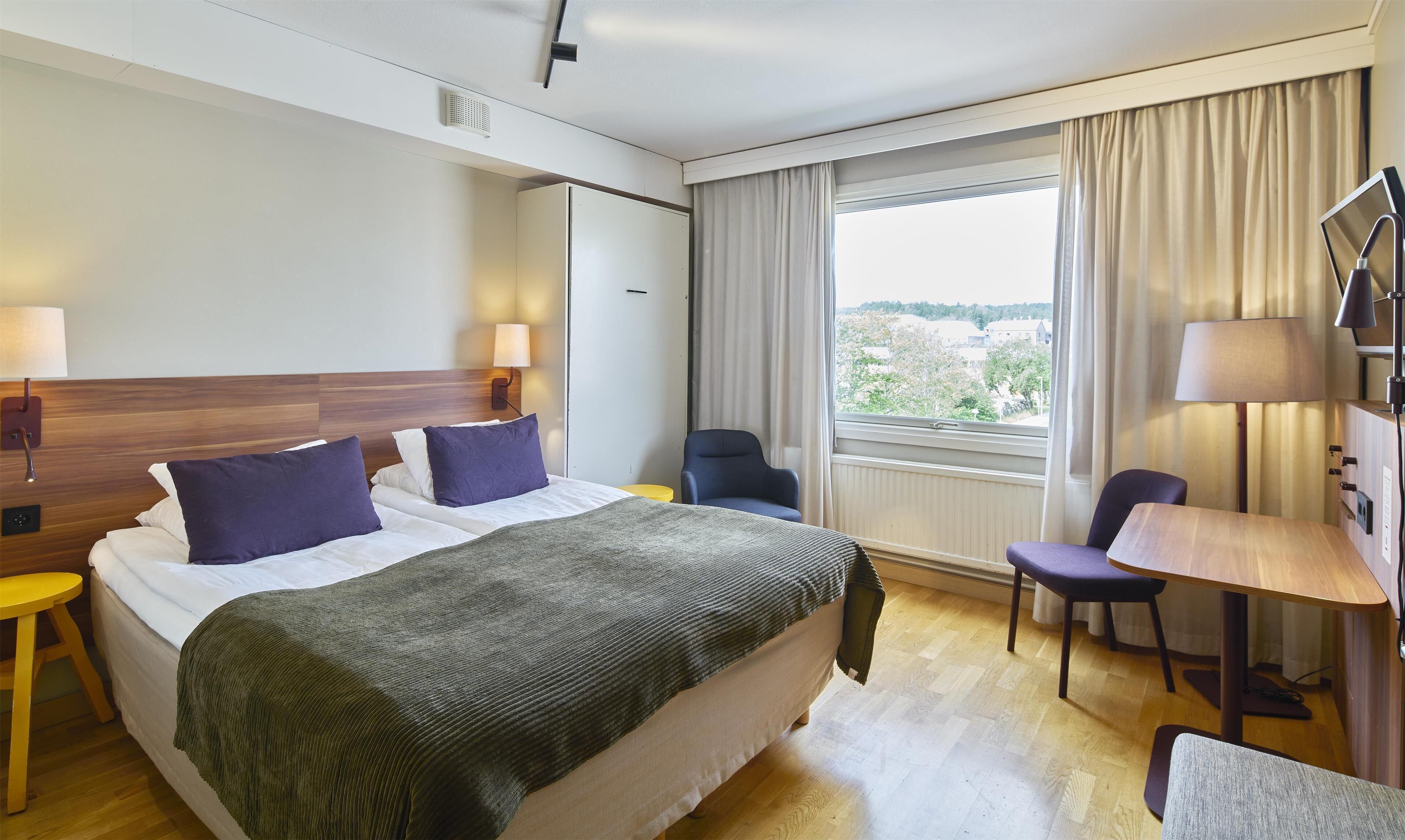 Quarto de hotel com uma cama grande, duas mesas de cabeceira, uma escrivaninha com cadeira, uma janela com cortinas e vista para o exterior.