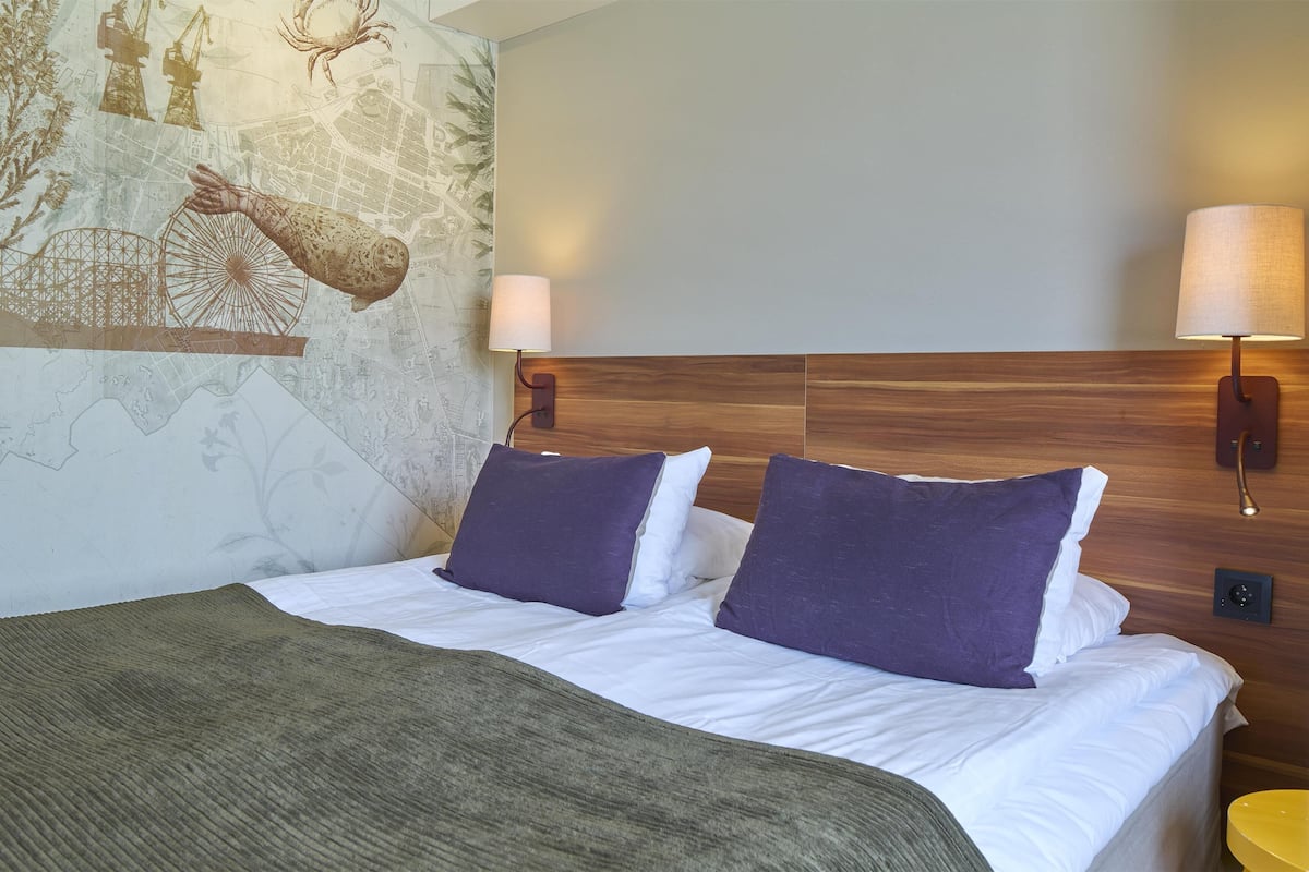 Ett hotellrum med en säng, två vägglampor, en träpanel vid sänggaveln och en dekorativ väggmålning.