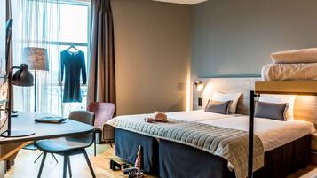 Quarto de hotel com cama, escrivaninha, luminária, cadeira e janela com cortinas.