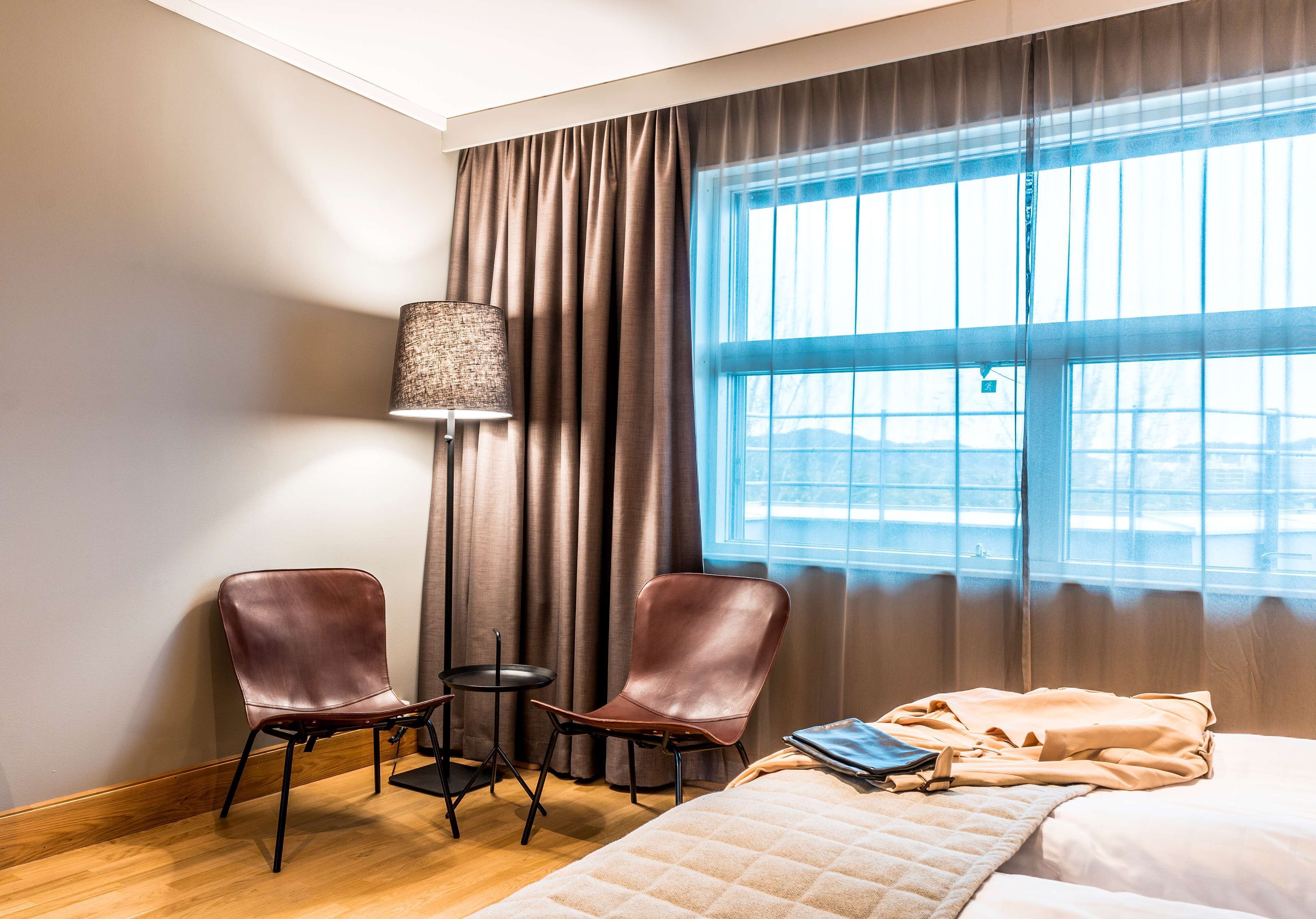 Une chambre d’hôtel avec un lit, deux fauteuils en cuir, une petite table, une lampe et une grande fenêtre avec des rideaux.
