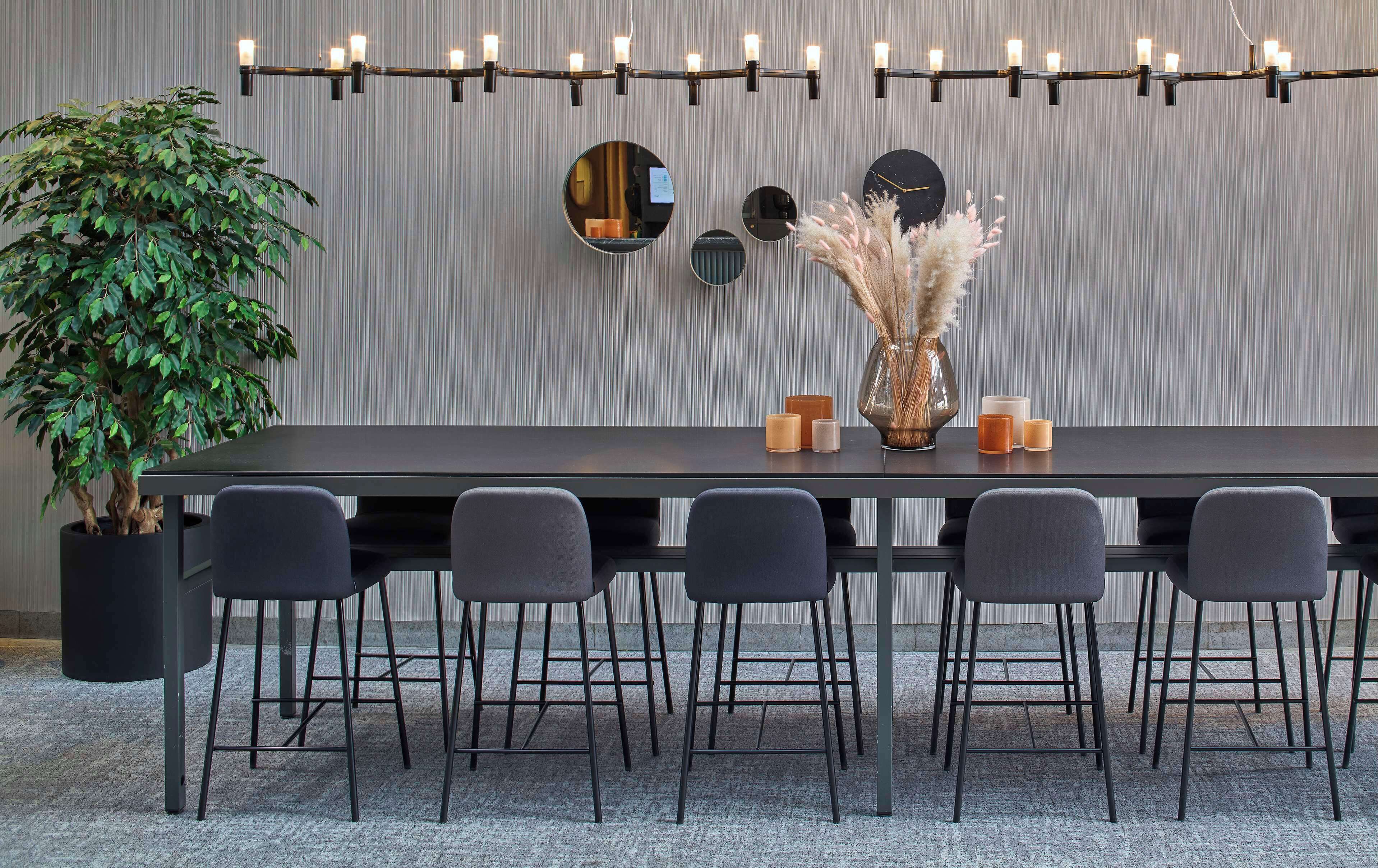 Un comedor moderno con una mesa larga, seis sillas, una planta en maceta, un jarrón con flores secas y una lámpara fijada a la pared.