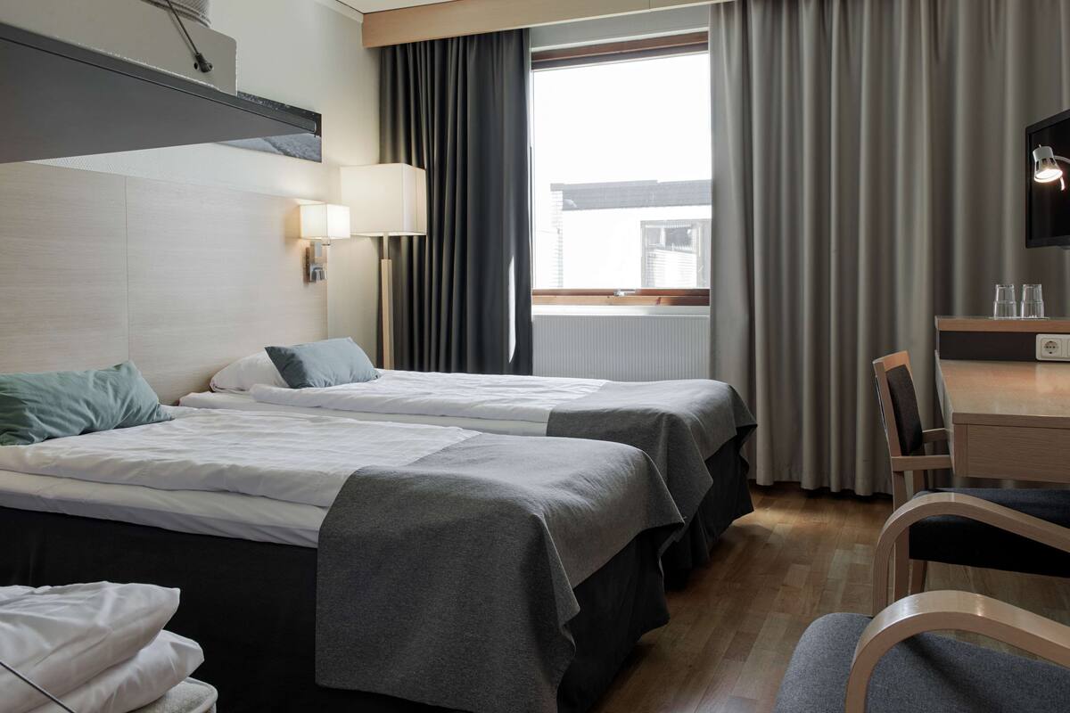 Habitación de hotel con dos camas, un escritorio, una silla, una lámpara y una ventana con cortinas.