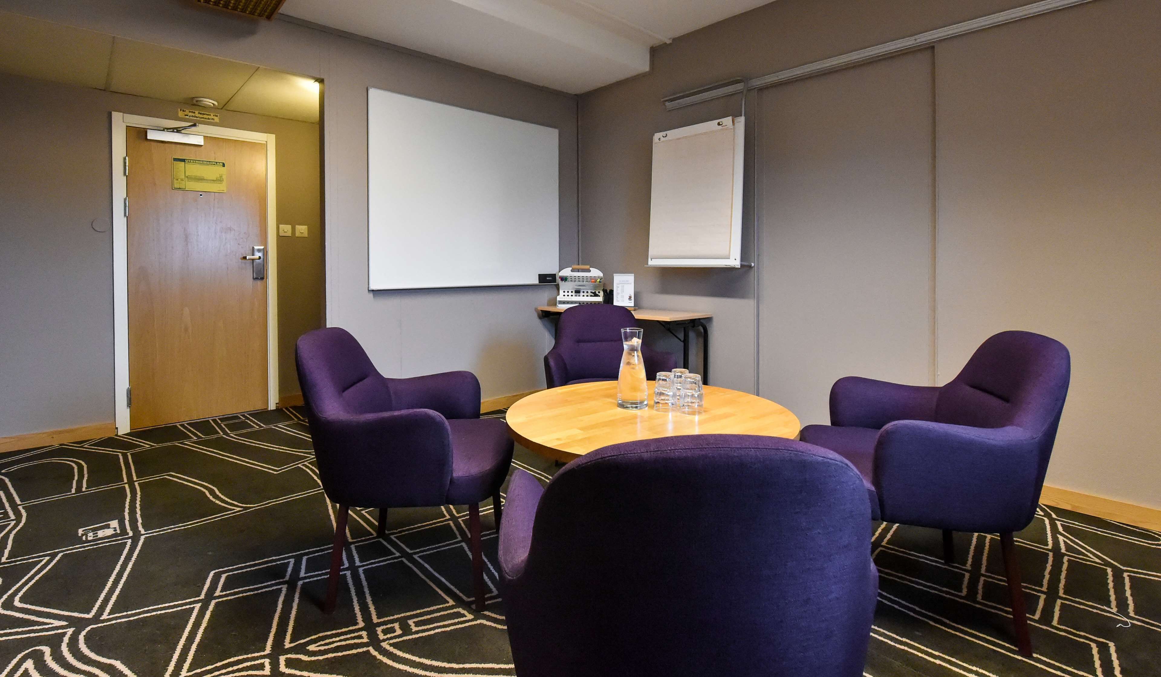 Una sala de conferencias con una mesa redonda, cuatro sillas moradas, una pantalla de proyector y una pizarra.