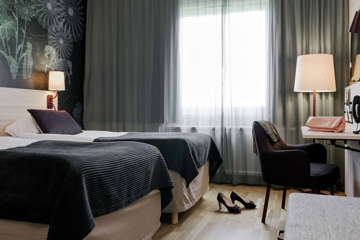 Habitación de hotel con dos camas, un escritorio con lámpara, una silla y un ventanal con cortinas.
