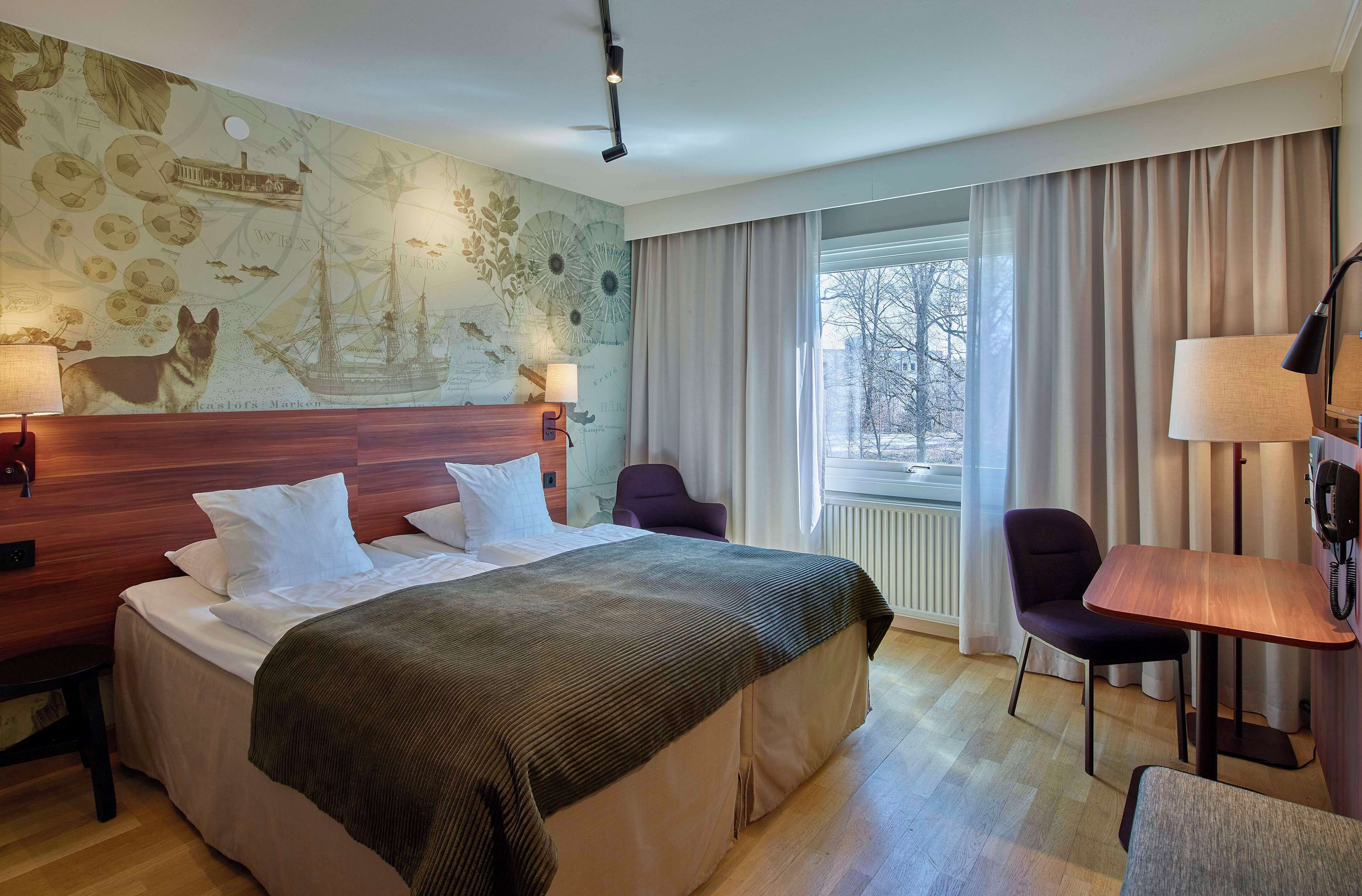 Quarto de hotel com uma cama grande, cabeceira de madeira, escrivaninha com cadeira, janela com cortinas e parede com mapa e ilustrações de navios.