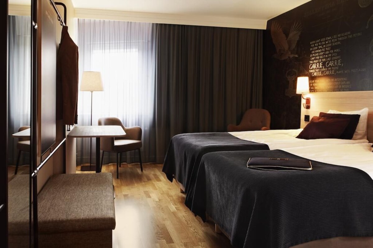 A hotel room with two beds, a desk, a chair, a lamp, and a large wall mural.