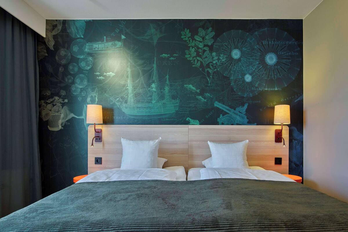 A hotel room with a large bed, two bedside lamps, and a wall mural with nautical and botanical elements.