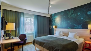 Habitación de hotel con una cama grande, un escritorio con silla, una mesita y una ventana con cortinas.