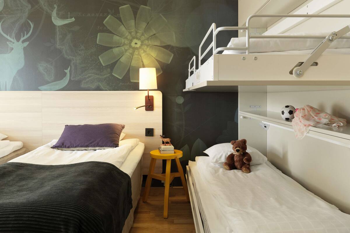 A hotel room with two beds, a small stool, a lamp, and a teddy bear.
