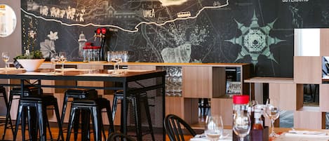 Un restaurant avec un mur noir orné d’une carte et d’un texte, des tables et des chaises en bois, et des luminaires suspendus.